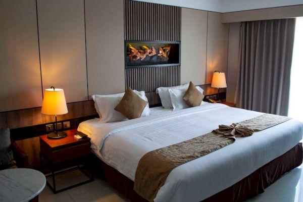 A neatly arranged hotel room features a large bed with pillows and blankets, warm lighting from table lamps, and a window with curtains.