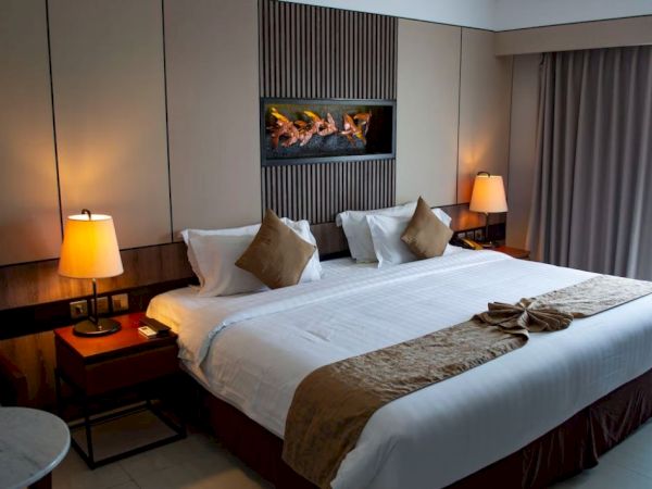 A neatly arranged hotel room features a large bed with pillows and blankets, warm lighting from table lamps, and a window with curtains.