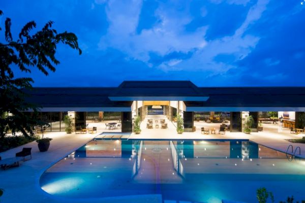 A luxurious modern building with a large, illuminated swimming pool in the foreground, captured at dusk with a beautiful blue sky overhead.