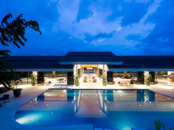 A luxurious modern building with a large, illuminated swimming pool in the foreground, captured at dusk with a beautiful blue sky overhead.