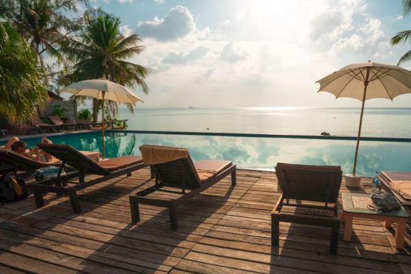 Poolside with lounge chairs, umbrellas, and palm trees overlooking a calm, scenic body of water under a bright sky with scattered clouds.
