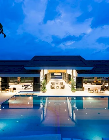 A luxurious building with an illuminated pool at dusk, featuring a spacious open area with seating arrangements and a clear blue sky.