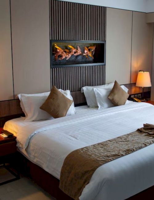 This image showcases a cozy hotel room with a king-sized bed, bedside tables with lamps, a wall-mounted fireplace, and a large window with curtains.