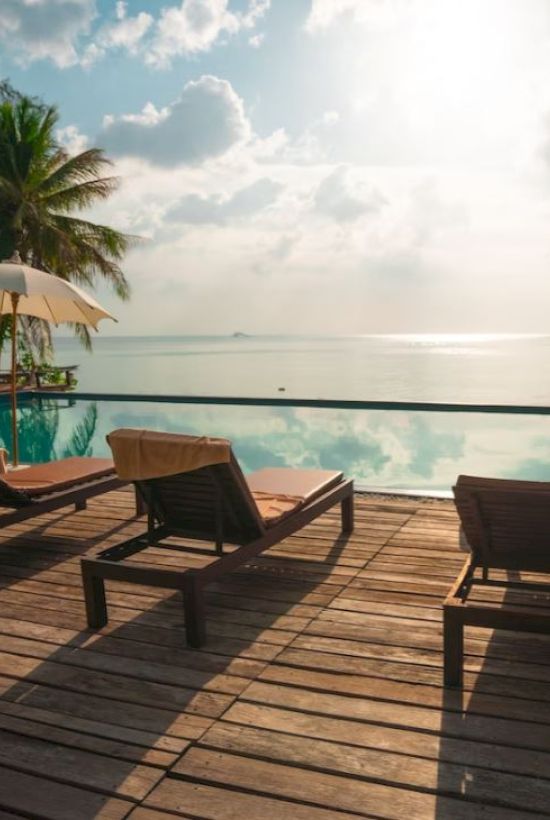 A serene poolside scene with lounge chairs, umbrellas, palm trees, and a view of the ocean under a bright, cloudy sky at sunset or sunrise.