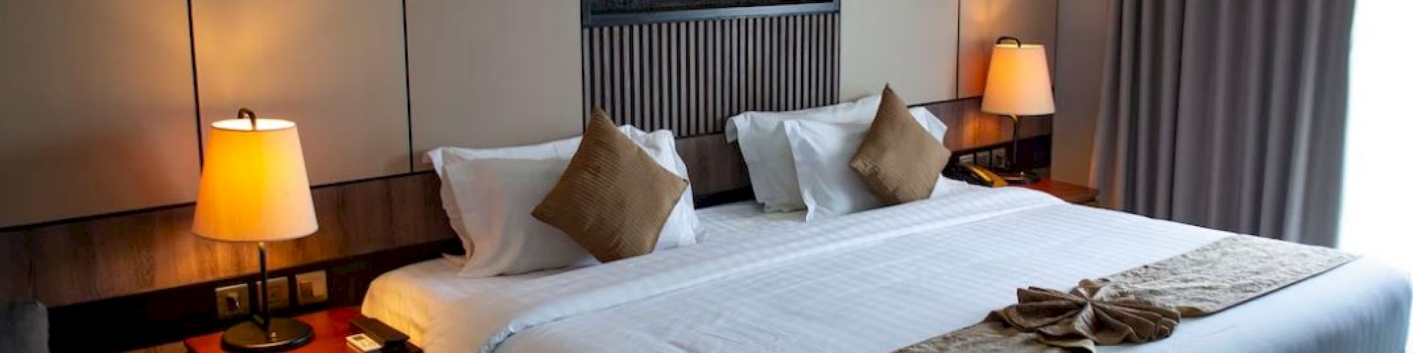 A well-maintained hotel room features a large, neatly made bed with pillows, bedside tables with lamps, a decorative feature on the wall, and curtains.