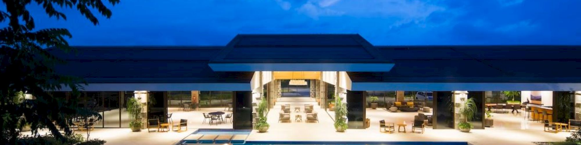 The image shows a luxurious outdoor pool area with lounge chairs and tables, adjacent to a modern building under a twilight sky.
