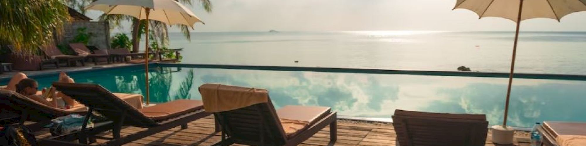 A serene poolside scene with lounge chairs, umbrellas, and a wooden deck overlooking a tranquil body of water under a partly cloudy sky with sunlight.