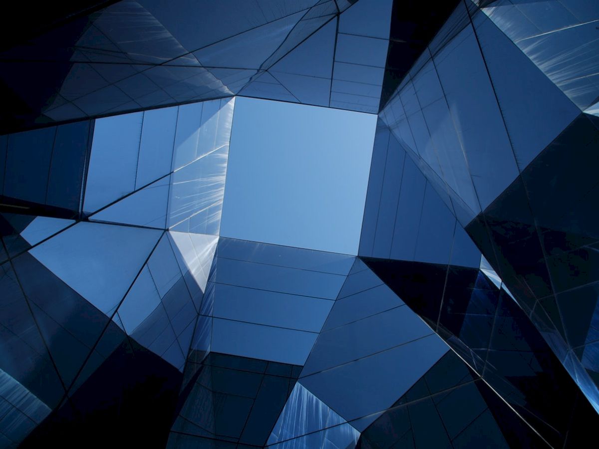 The image shows a geometric architectural structure with reflective glass surfaces forming a symmetrical pattern against a bright blue sky.