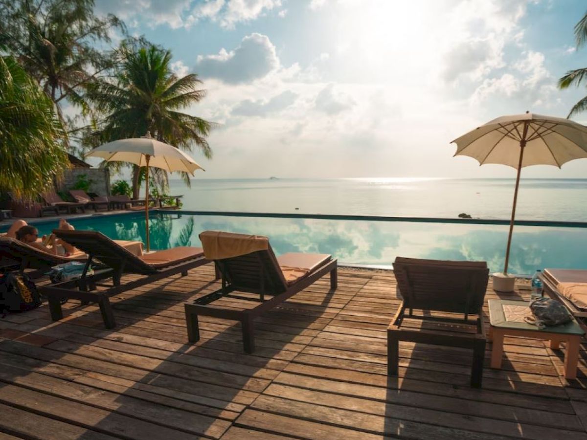 A tropical poolside scene with lounge chairs, umbrellas, palm trees, and a serene view of the ocean under a partly cloudy sky.