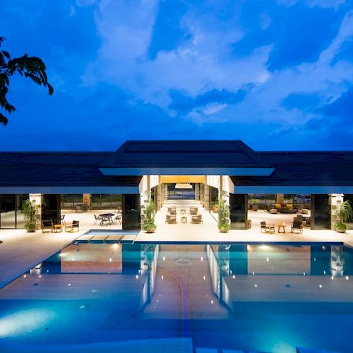 A modern resort or luxury residence with a large illuminated pool at dusk, surrounded by lounge chairs, and a spacious, well-lit interior.