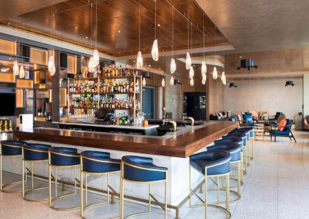 This image shows a modern bar with a long counter, blue bar stools, hanging lights, and shelves stocked with various bottles.