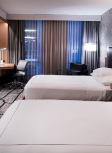 The image shows a modern hotel room with two beds, a desk, a television, and a window with curtains, creating a cozy and functional atmosphere.