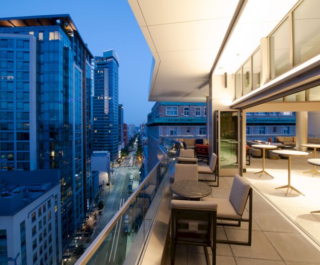 The image shows a modern outdoor terrace with seating, overlooking a cityscape of tall buildings under a dusky sky.