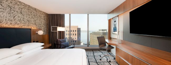 Modern hotel room with a large bed, a TV on a wooden wall unit, a desk with a chair, a lounge chair, and a large window with a city view.