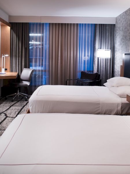 A modern hotel room features two neatly made beds, a flat-screen TV, a desk with a chair, and contemporary decor with ample lighting.