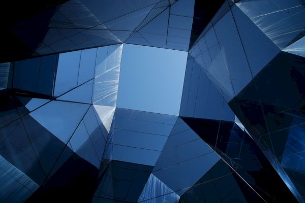 The image shows an abstract architectural design with mirrored glass panels forming geometric shapes, reflecting the sky in a modern, faceted pattern.