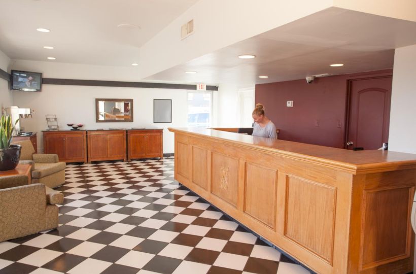 A hotel lobby with a checkered floor, wooden reception desk, a person behind it, seating area, wall art, TV, and decorative plants, ending the sentence.