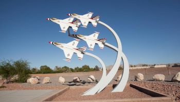 A statue of four fighter jets in an aerial formation, displayed on curved pillars, in an outdoor setting with rocks and sparse vegetation.