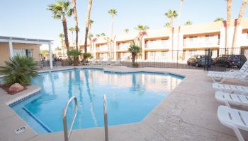 The image shows a swimming pool surrounded by lounge chairs and palm trees, with a two-story building and parked cars in the background.