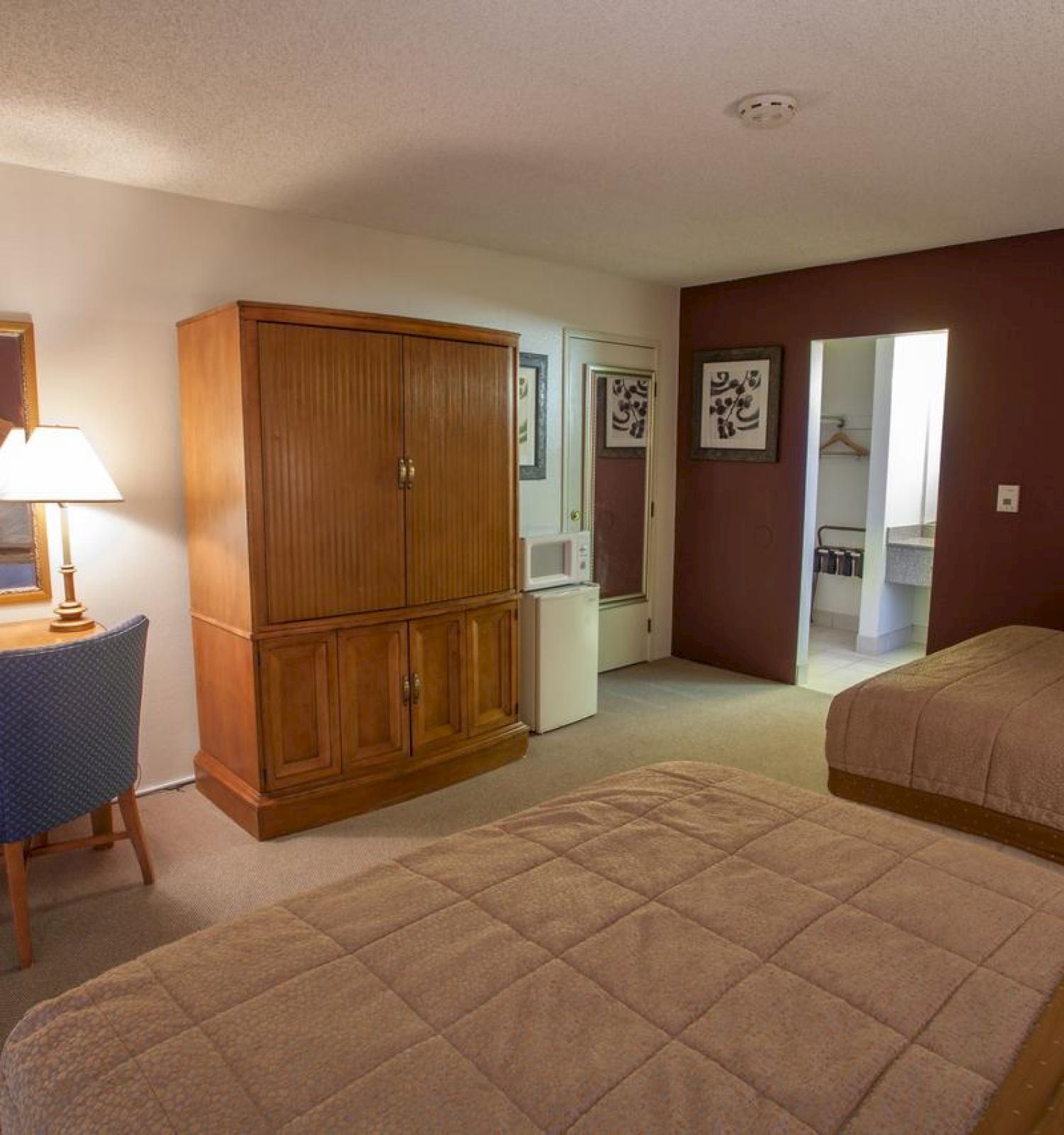 This image shows a hotel room with two beds, a wooden wardrobe, a desk with a mirror and chair, a lamp, and framed art on the walls.