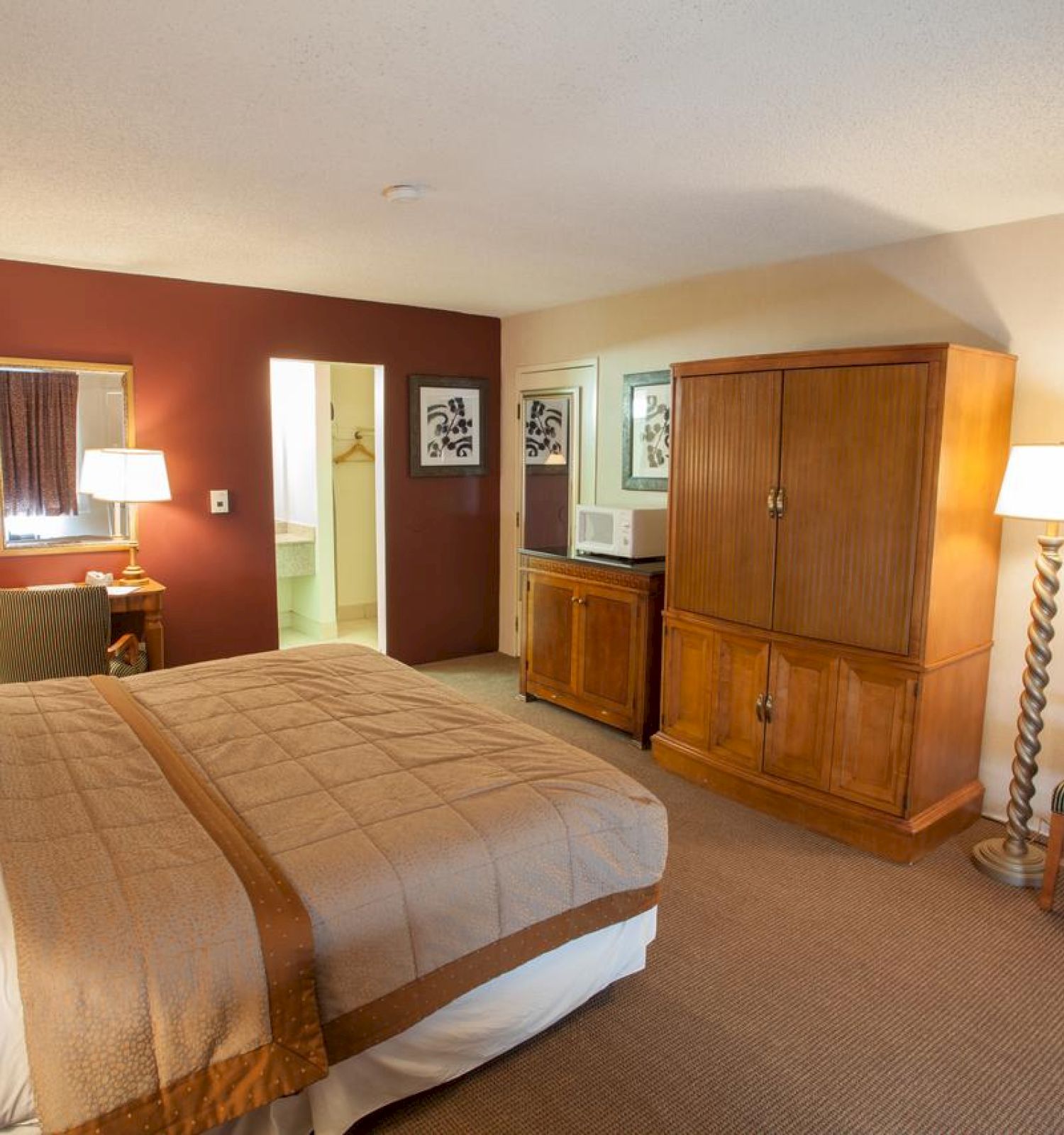 A cozy hotel room with a bed, wooden furniture, a microwave, lamps, a chair, framed art, and a bathroom door partially open.