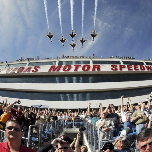 A crowd at a race track watches as jets fly over in formation, leaving contrails in the sky above the 