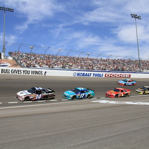 This image shows a group of race cars speeding on a track, with spectators in the background and advertisements displayed on the track walls.