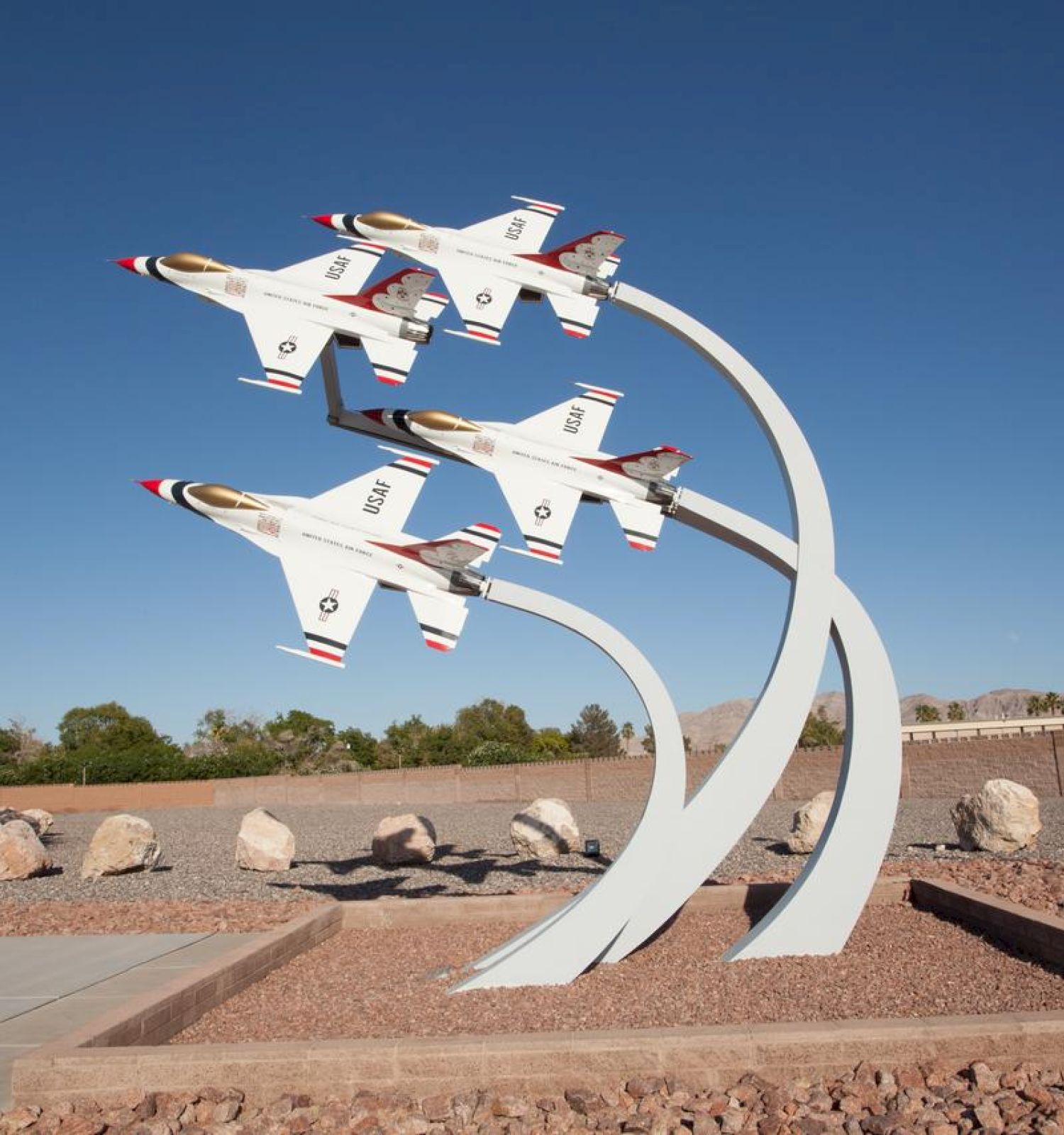 A sculpture of four fighter jets in formation, mounted on curved supports, is displayed outdoors in a desert landscape with rocks and plants, under a clear sky.