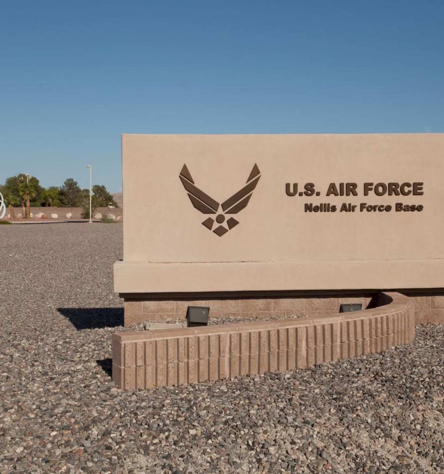 A signboard for the U.S. Air Force, Nellis Air Force Base, set in a gravel landscape with trees and structures in the background.