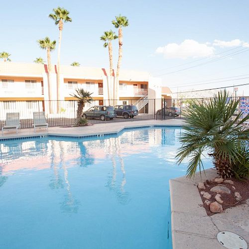 The image depicts an outdoor pool surrounded by lounge chairs and palm trees with a motel-like building and parked cars in the background.