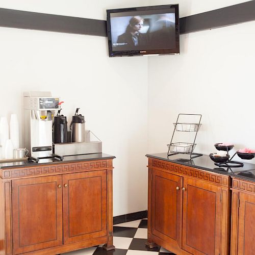 The image shows a coffee station with coffee dispensers, cups, and snacks, along with a TV mounted on the wall in a room with checkered flooring.