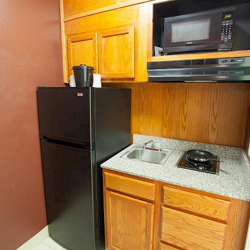 A compact kitchenette with a black fridge, wooden cabinets, a microwave, a sink, and a stovetop on a granite countertop.