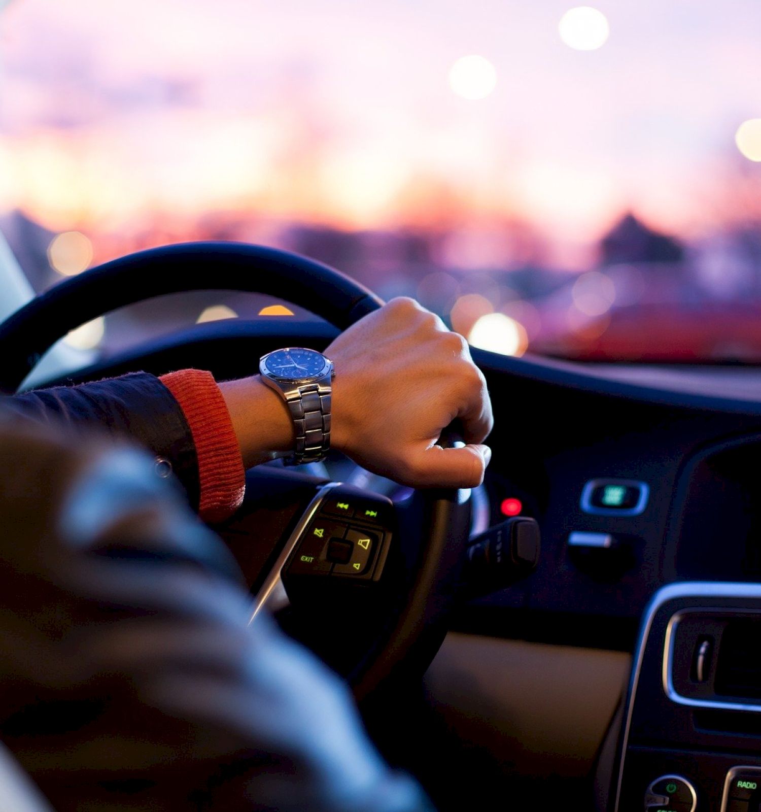 A person is driving a car at dusk, with one hand on the steering wheel and the dashboard illuminated with lights and gauges.