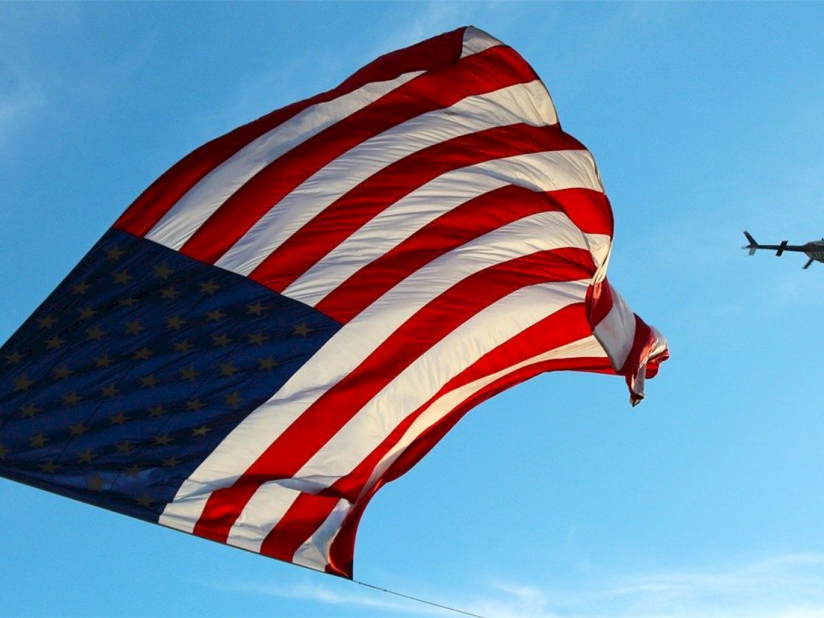 A large American flag is waving in the sky on a sunny day with a helicopter flying in the background.