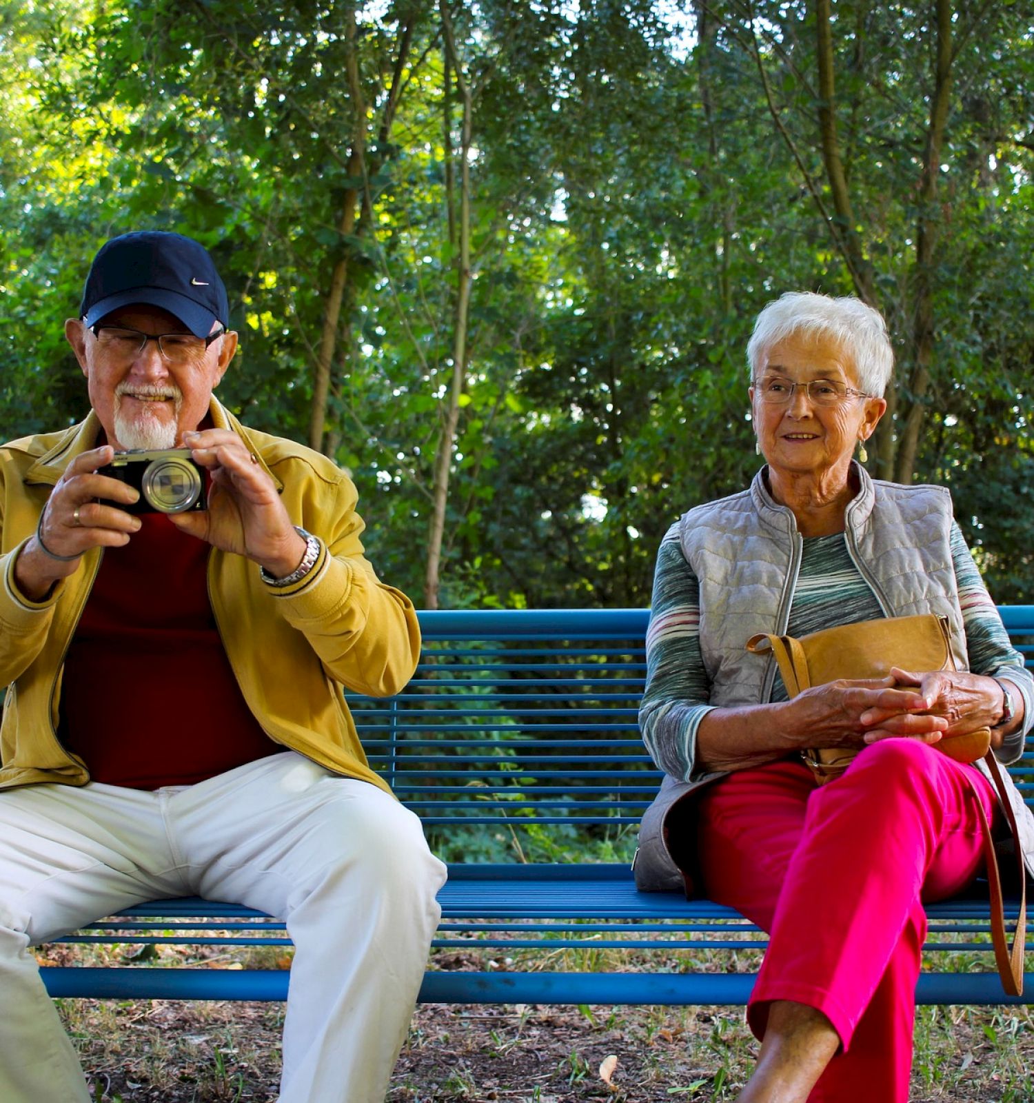 An elderly man and woman sit on a bench in a wooded area; he is holding a camera, and she is looking ahead, smiling.