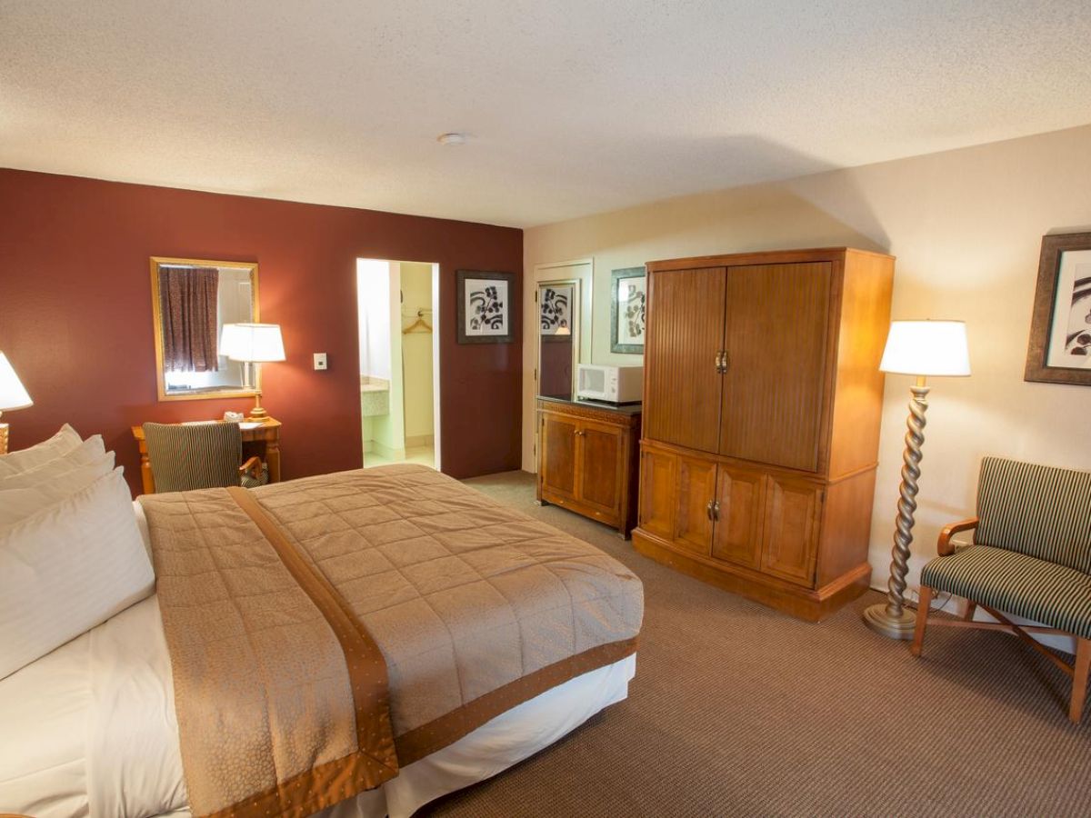 A cozy hotel room with a double bed, wooden furniture, a lamp, chair, TV, microwave, and a door leading to a bathroom at the far end.