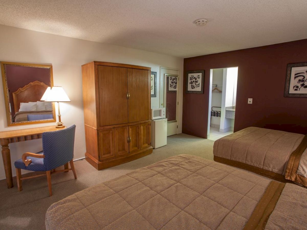 This image shows a hotel room with two beds, a wooden wardrobe, a desk with a mirror and lamp, and an open door to a bathroom area.