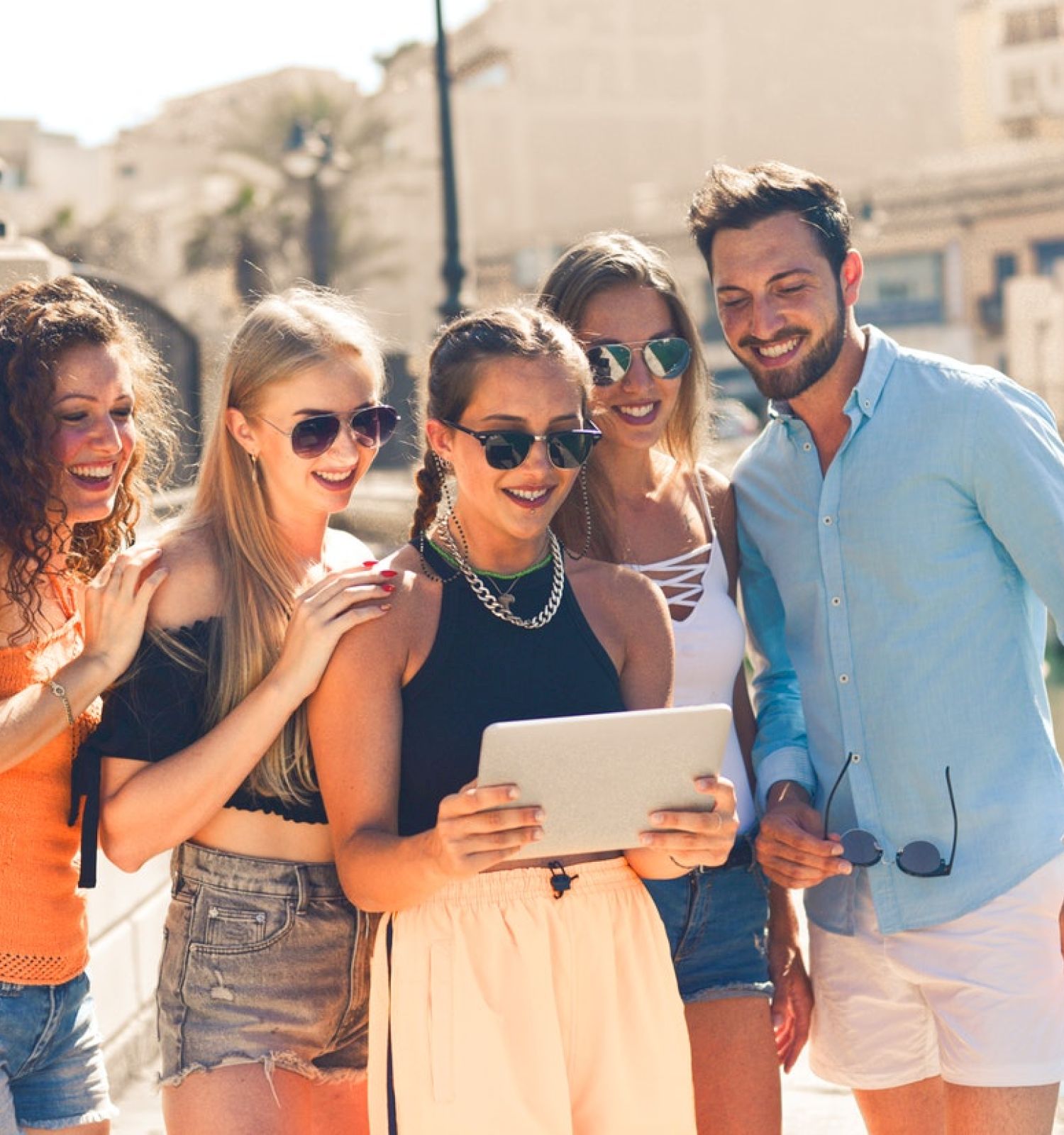 A group of five people, three women and two men, are enjoying themselves outdoors, gathered around a tablet, smiling and seeming excited.