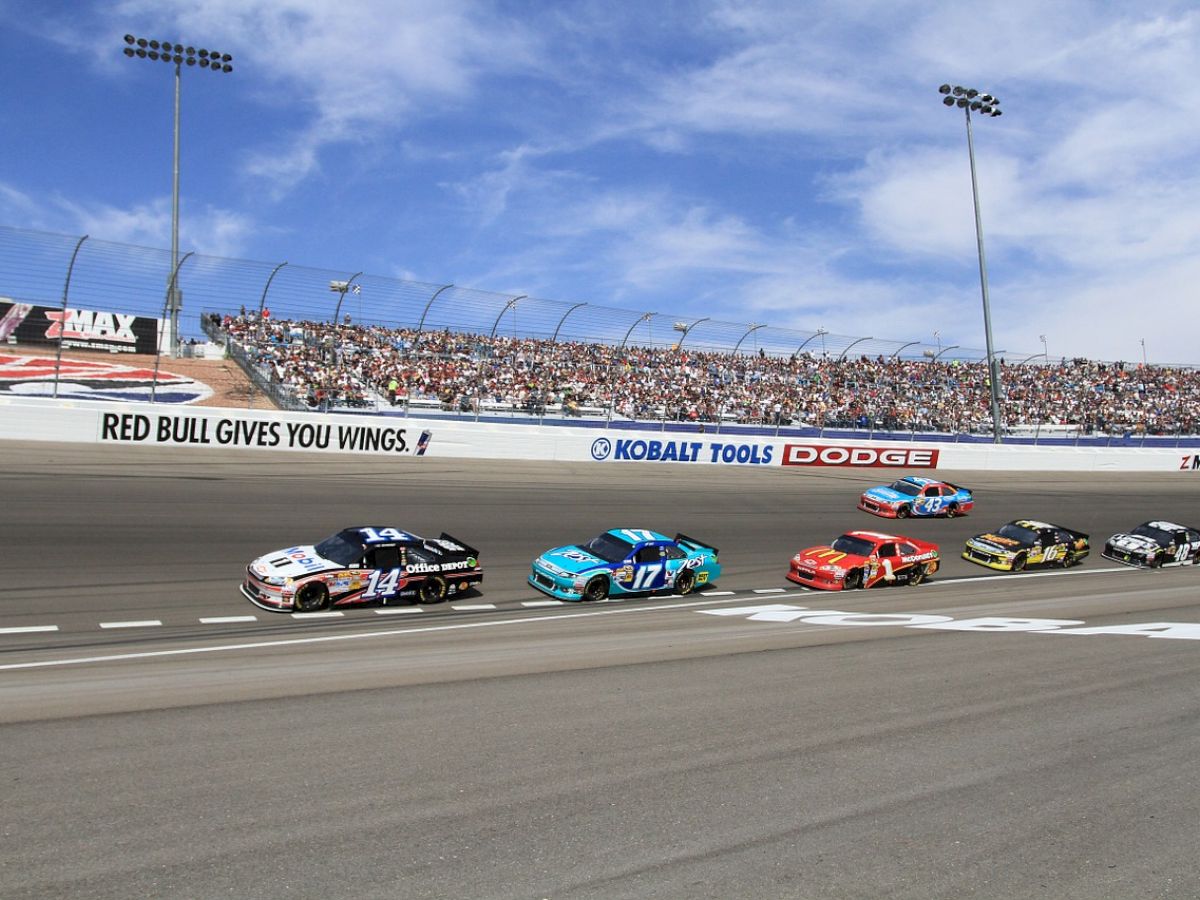 A group of race cars competing on a track with a crowd of spectators in the stands and advertisements displayed, under a partly cloudy sky.