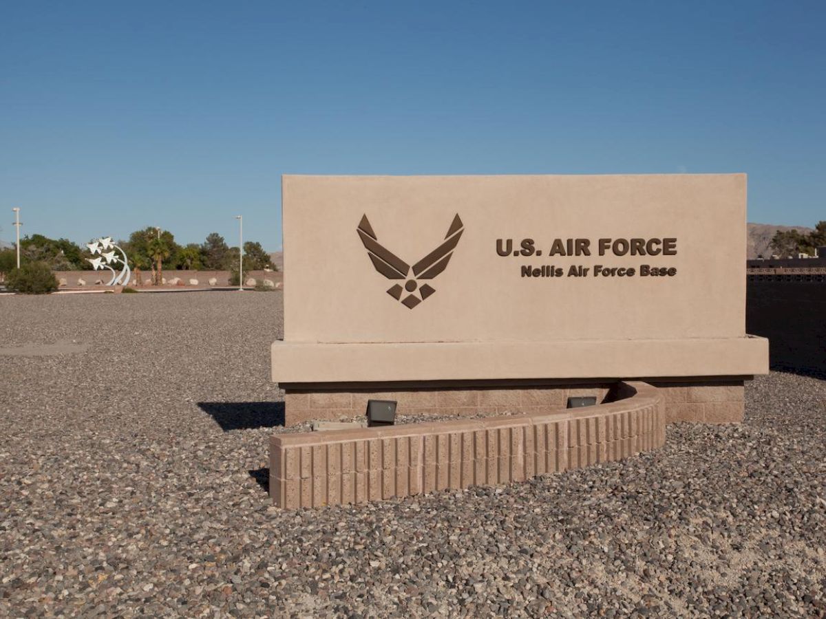 A sign for Nellis Air Force Base with the U.S. Air Force logo, situated in a gravel-covered area.