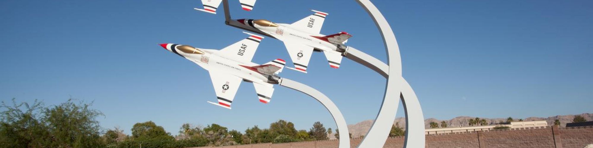 The image shows a sculpture of four fighter jets arranged to appear in flight, against a clear blue sky, with desert landscaping in the background.