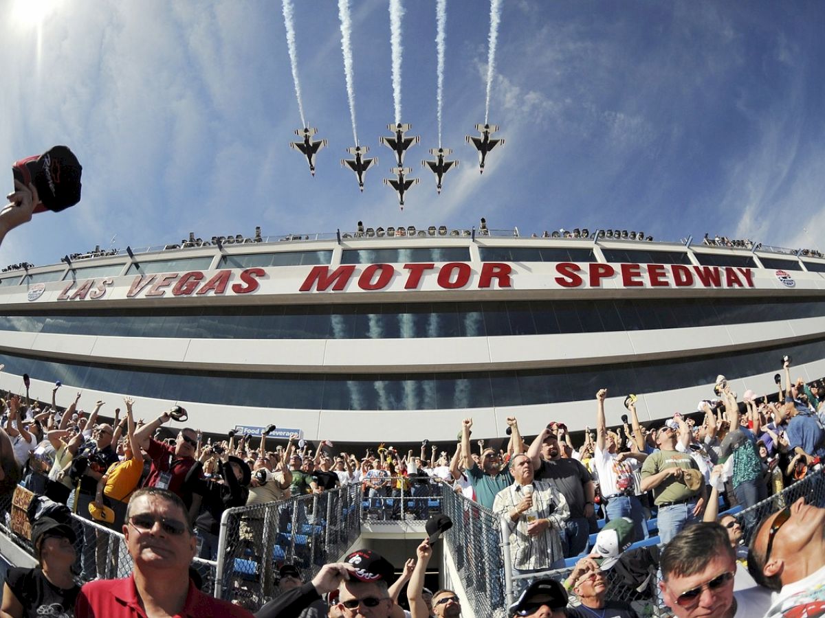 A crowd at Vegas Motor Speedway watches a formation of jets flying overhead, leaving white trails against a clear blue sky.