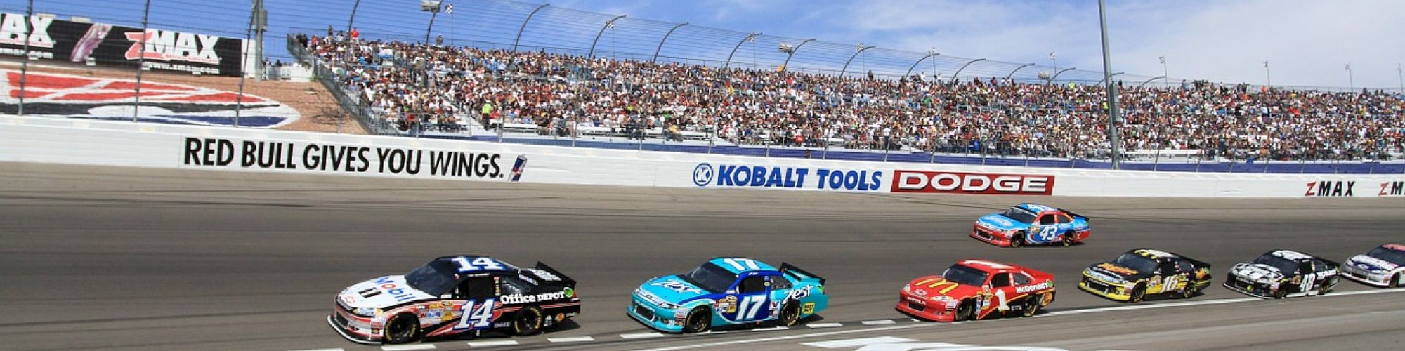 A group of race cars is competing on a track with spectators in the stands and advertisements visible on the barriers.