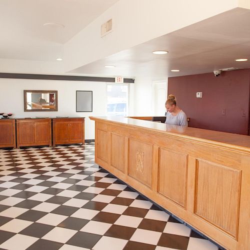 The image depicts a hotel lobby with checkered flooring, a wooden front desk, a person behind the desk, seating area, and a TV on the wall.