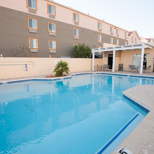 An outdoor pool with clear blue water is surrounded by a concrete deck, lounge chairs, and a covered seating area, adjacent to a multi-story building.