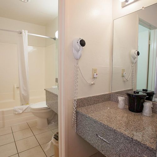 The image shows a bathroom with a bathtub and shower, toilet, granite countertop with a sink, hairdryer, and mirror. The floor is tiled.