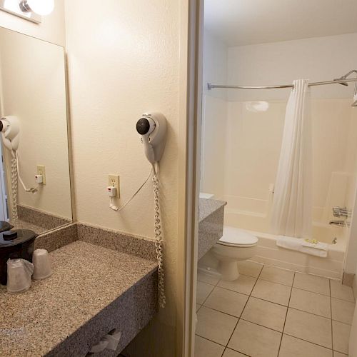 The image shows a bathroom with a granite countertop, mirror, hairdryer, and sink in the foreground. A bathtub with a shower curtain is in the background.