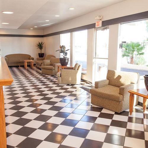 A spacious lobby with checkered flooring, several seating areas, large windows, and indoor plants.