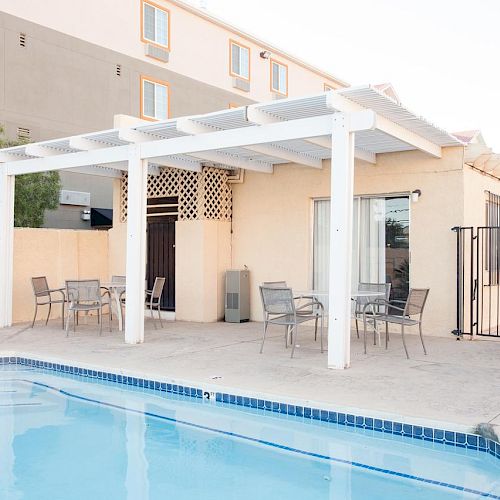 The image shows an outdoor pool area with a shaded seating area, tables, and chairs next to a building in a bright, sunny setting.