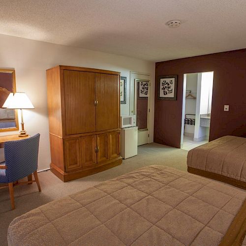 This image shows a hotel room with two beds, a wooden closet, a desk with a lamp and mirror, and a bathroom in the background.